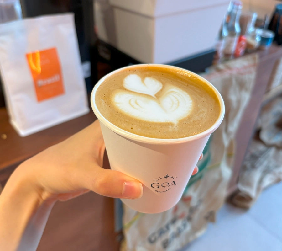 Hand holding a cup of coffee with milk, featuring heart-shaped latte art, served in a Goa Specialty Coffee branded cup.